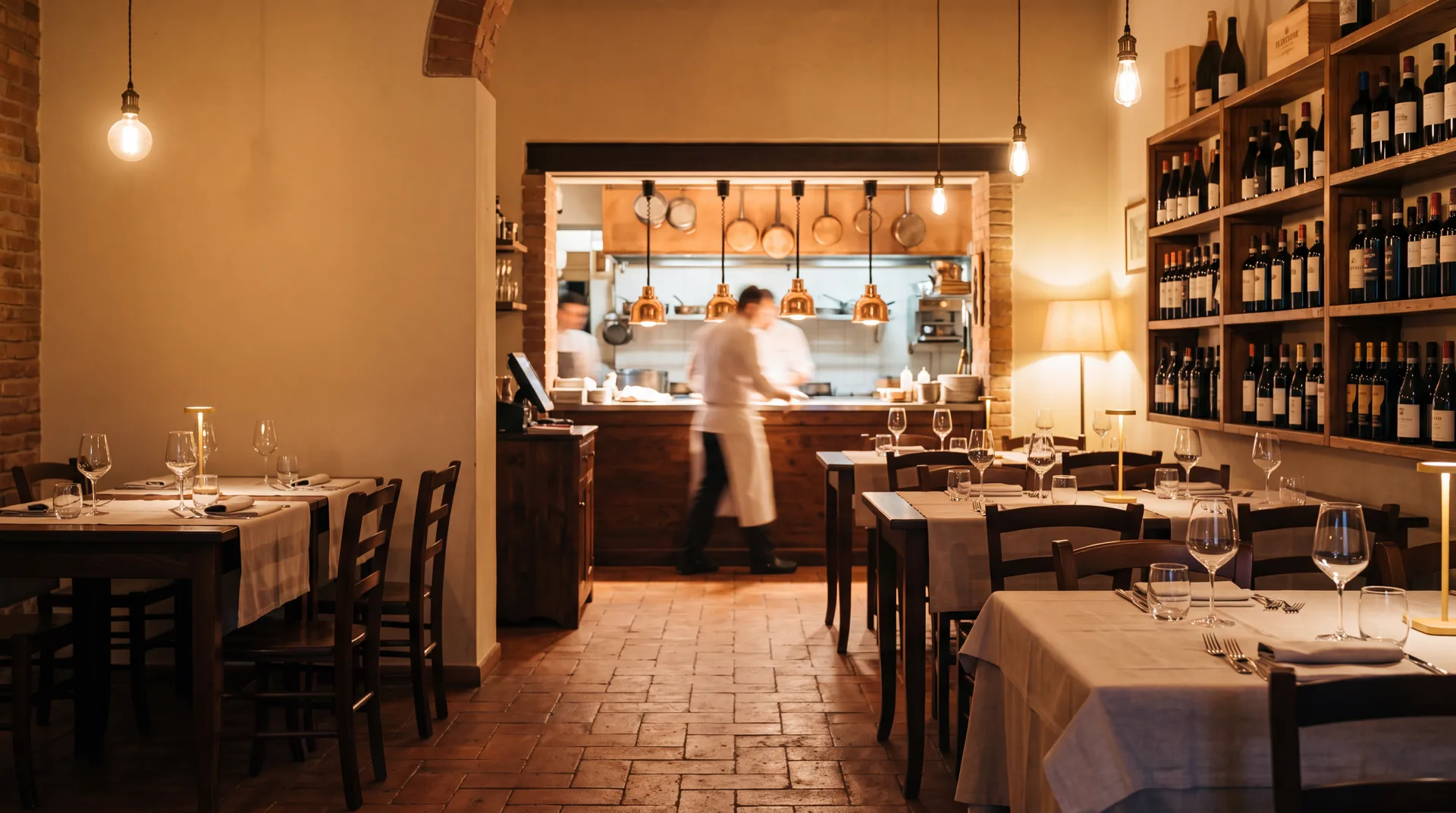 Sala di un ristorante italiano nell'ora prima del servizio, tavoli apparecchiati e cucina illuminata sul fondo